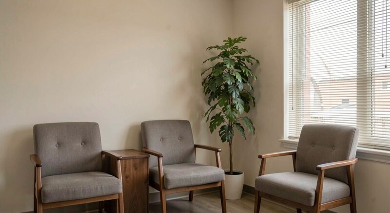 A quiet waiting room corner with three upholstered chairs and a tall green plant, plain wall, soft daylight through blinds